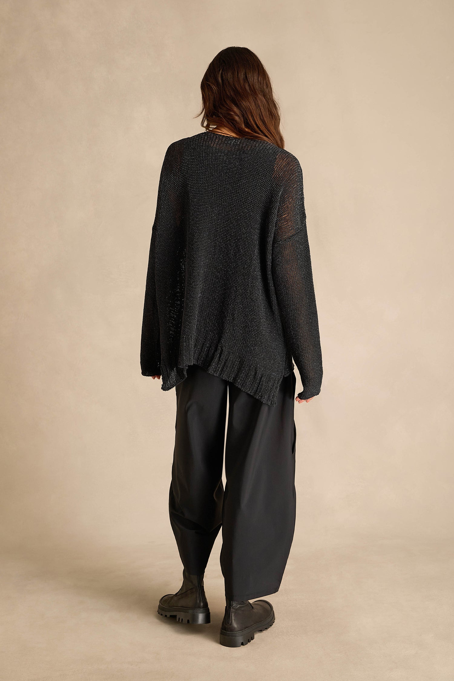 A person stands with their back to the camera, wearing the Black Seam Detail Scoop Neck Knit, paired with wide black pants and black boots, set against a beige background.