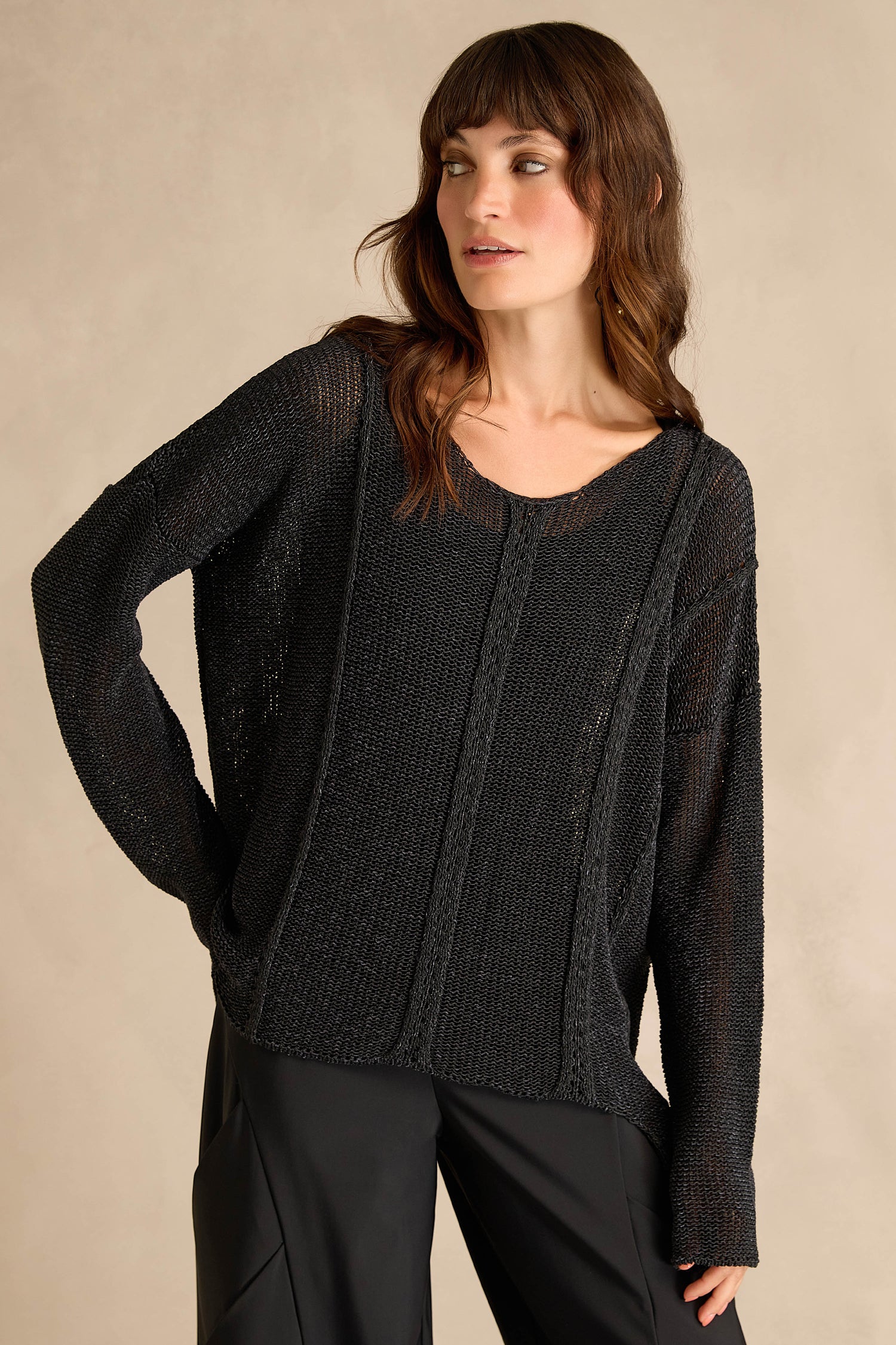 A woman wearing the Black Seam Detail Scoop Neck Knit, a cotton-rich longline black sweater with long sleeves, paired with black pants, stands against a neutral beige background.