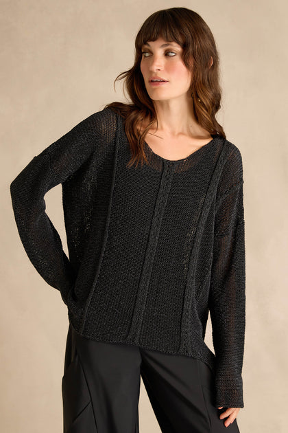 A woman wearing the Black Seam Detail Scoop Neck Knit, a cotton-rich longline black sweater with long sleeves, paired with black pants, stands against a neutral beige background.