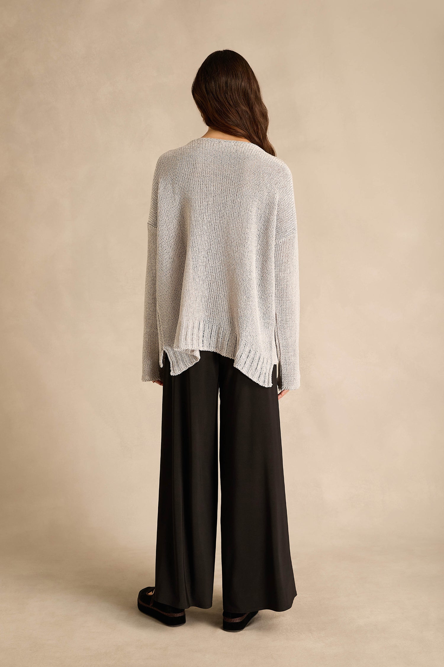 A person stands with their back to the camera, wearing the Silver Seam Detail Scoop Neck Knit in light gray and wide-leg black pants, set against a neutral beige background.
