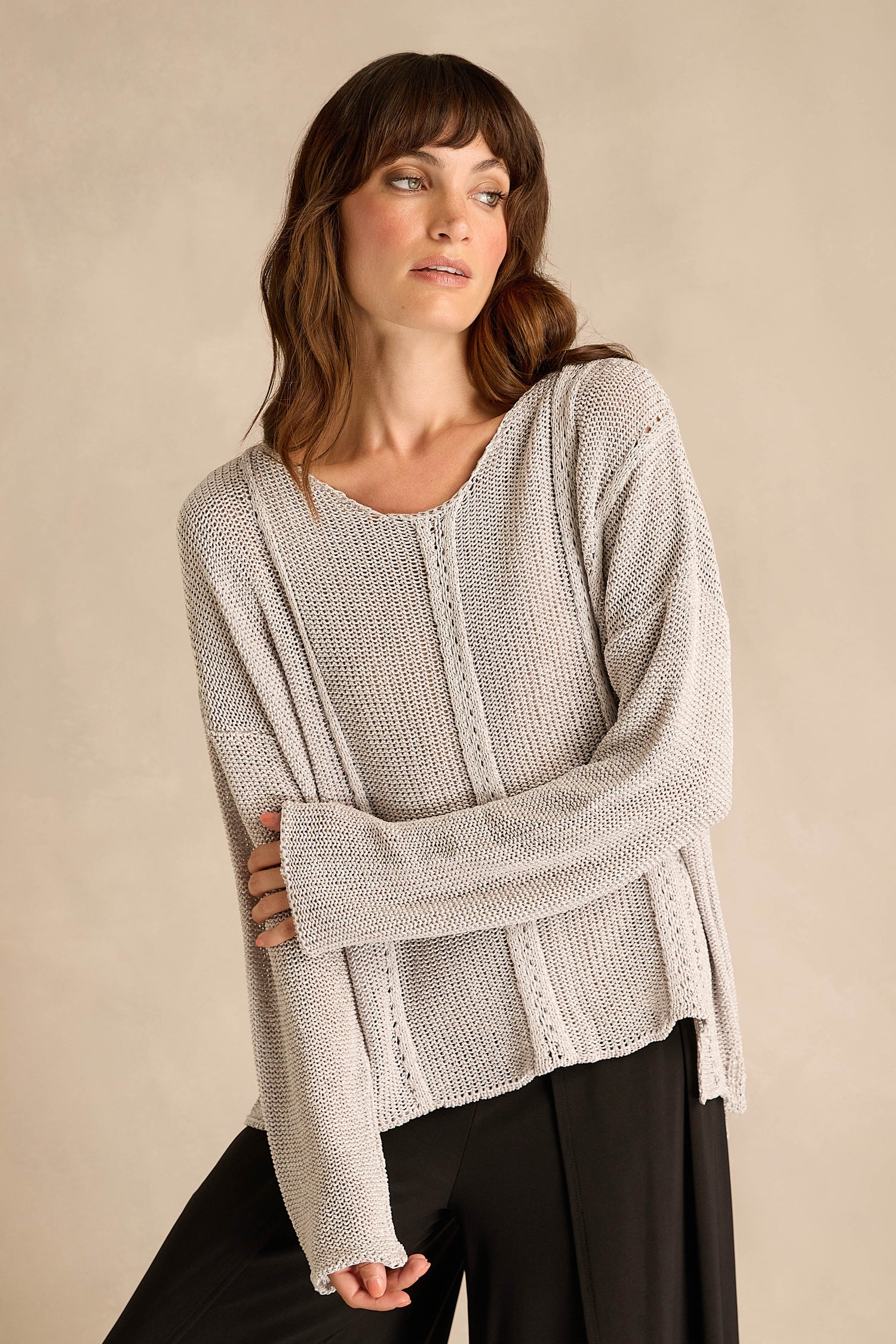 A woman wearing the Silver Seam Detail Scoop Neck Knit, a loose, light beige cotton-rich sweater featuring front seam detailing, pairs it with black pants and stands against a plain beige background, looking to the side.