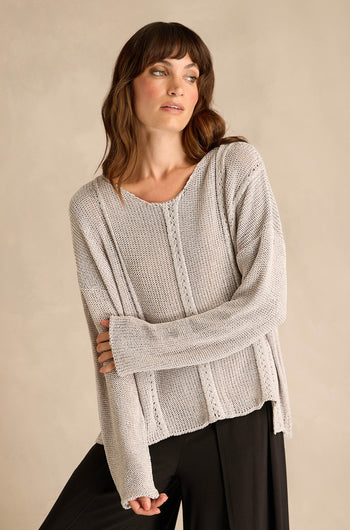 A woman wearing the Silver Seam Detail Scoop Neck Knit, a loose, light beige cotton-rich sweater featuring front seam detailing, pairs it with black pants and stands against a plain beige background, looking to the side.
