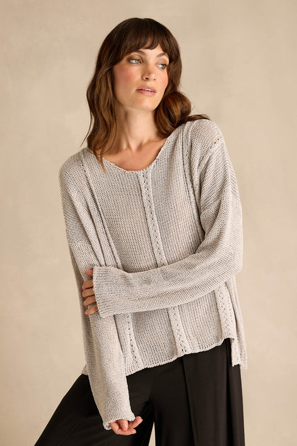 A woman wearing the Silver Seam Detail Scoop Neck Knit, a loose, light beige cotton-rich sweater featuring front seam detailing, pairs it with black pants and stands against a plain beige background, looking to the side.