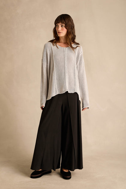A woman stands against a plain beige background, looking to the side, wearing the Silver Seam Detail Scoop Neck Knit sweater in light gray paired with wide-leg black pants.
