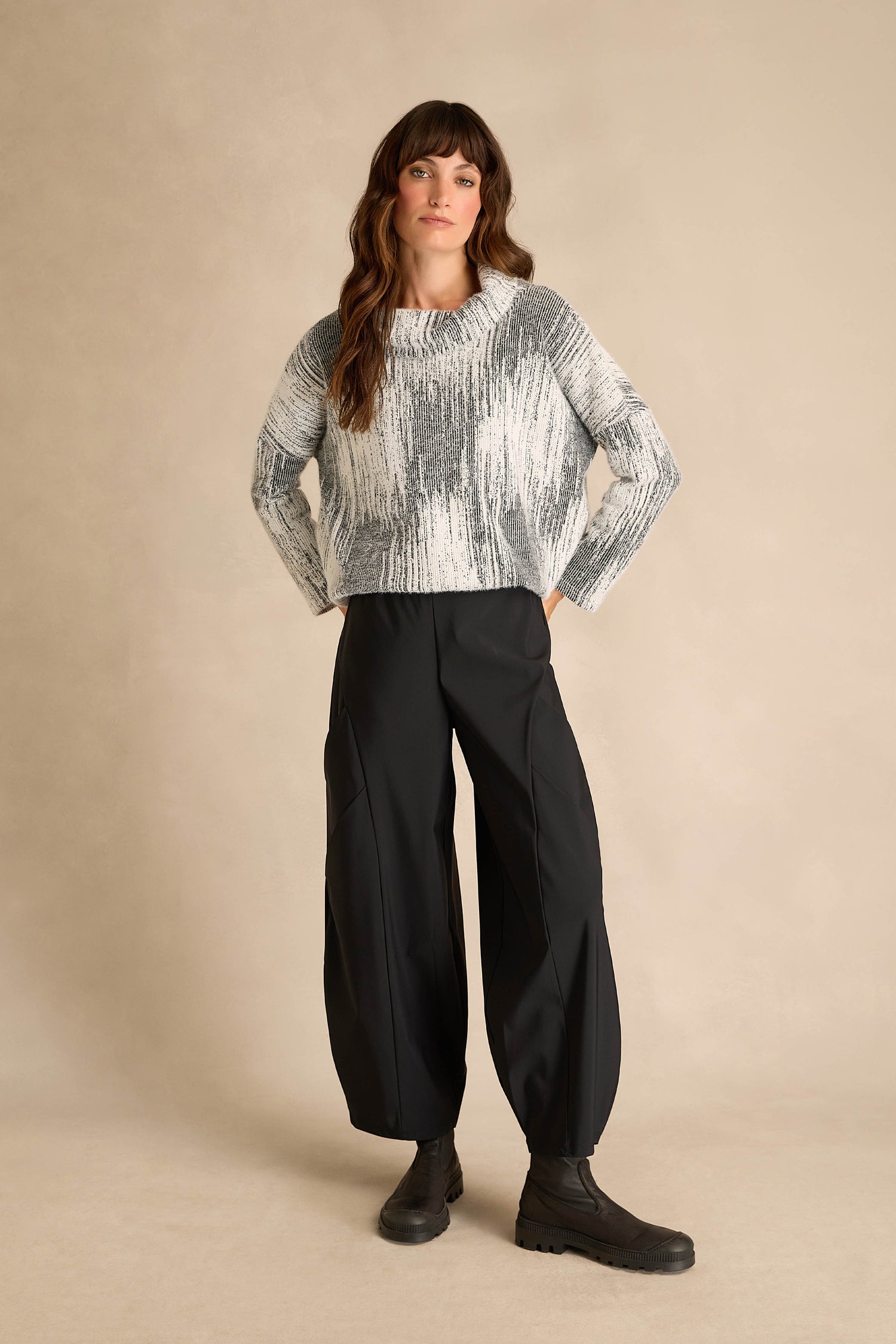 Person standing against a beige background, wearing a gray and white knit sweater, Seamed Bubble Trousers in black, and black boots.
