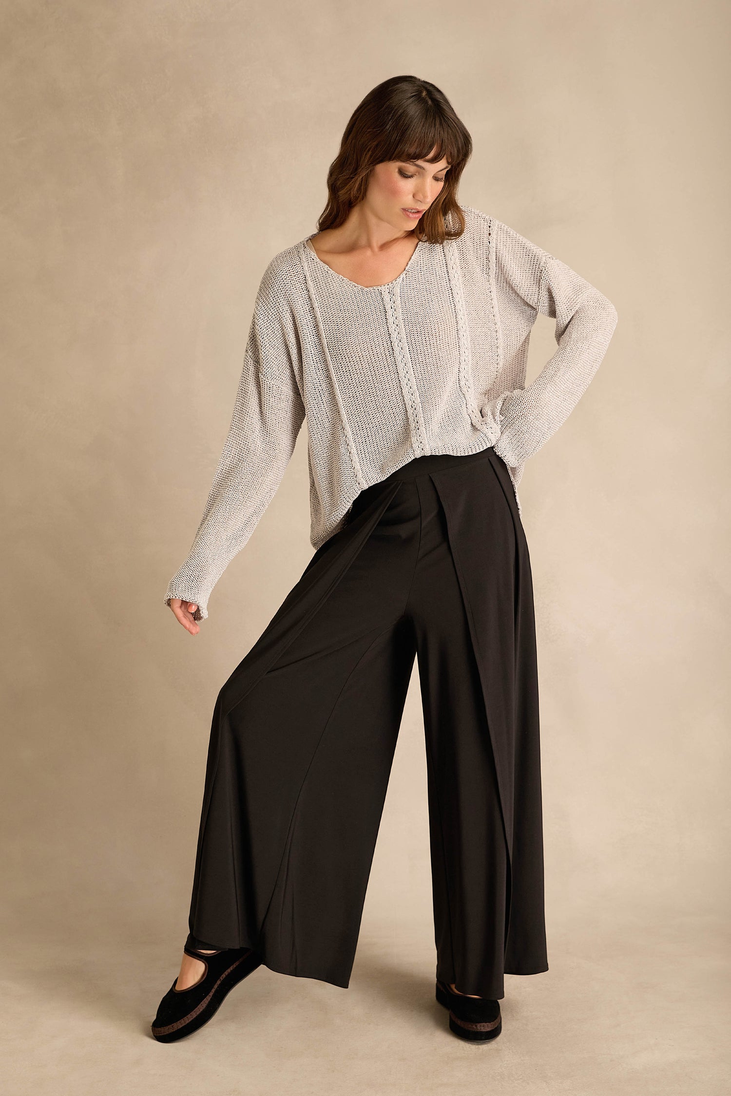 Woman wearing a loose knit sweater and Wide Leg Wrap Jersey Trousers stands with one hand on her hip and looks down against a neutral background.