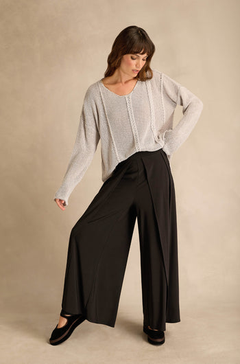 Woman wearing a loose knit sweater and Wide Leg Wrap Jersey Trousers stands with one hand on her hip and looks down against a neutral background.