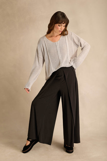 Woman wearing a loose knit sweater and Wide Leg Wrap Jersey Trousers stands with one hand on her hip and looks down against a neutral background.