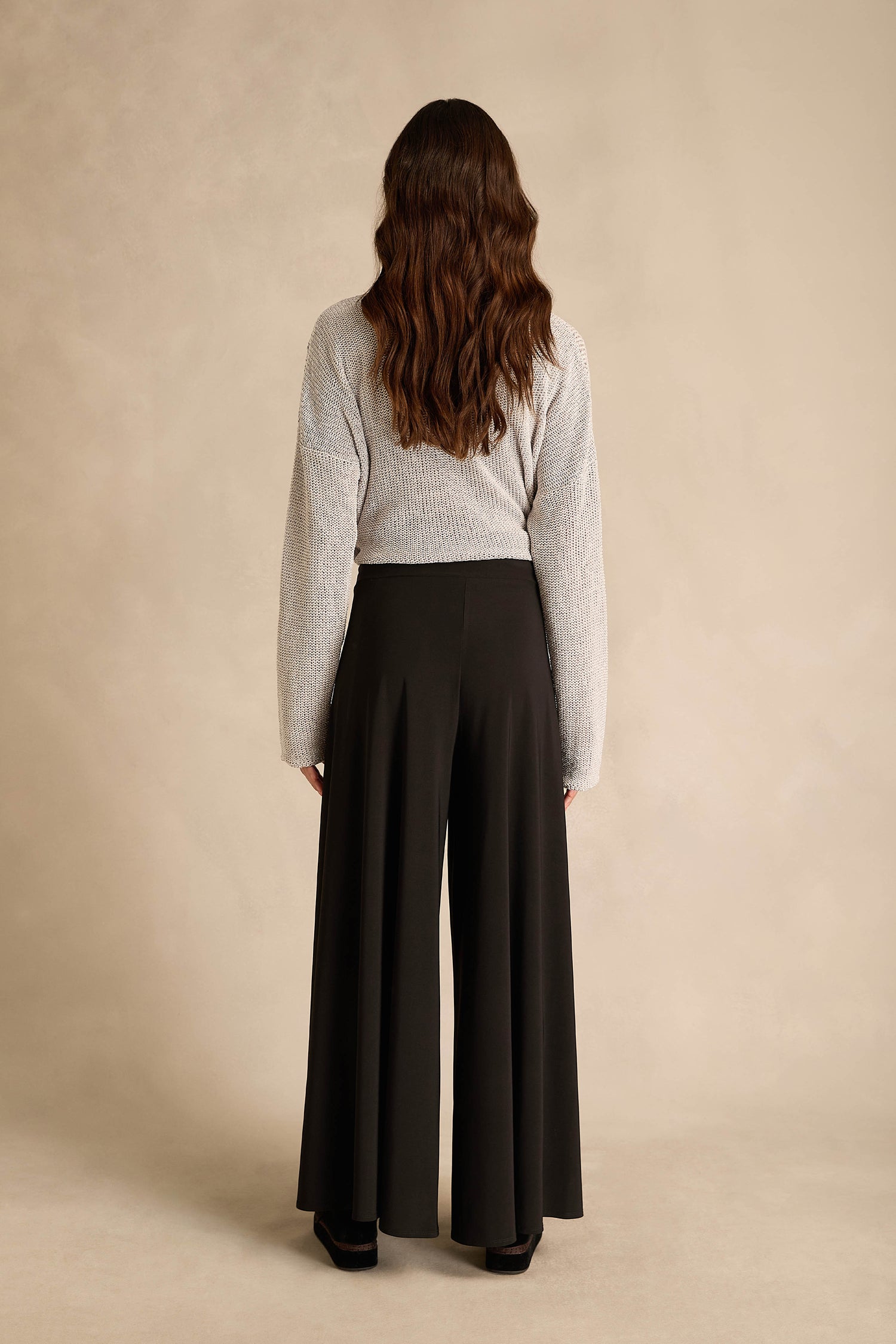 A person with long wavy brown hair, wearing a gray sweater and the Wide Leg Wrap Jersey Trousers, stands facing away from the camera against a beige background.