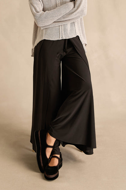 A person wearing a light knit sweater, Wide Leg Wrap Jersey Trousers, and black platform Mary Jane shoes stands with legs crossed against a neutral background.