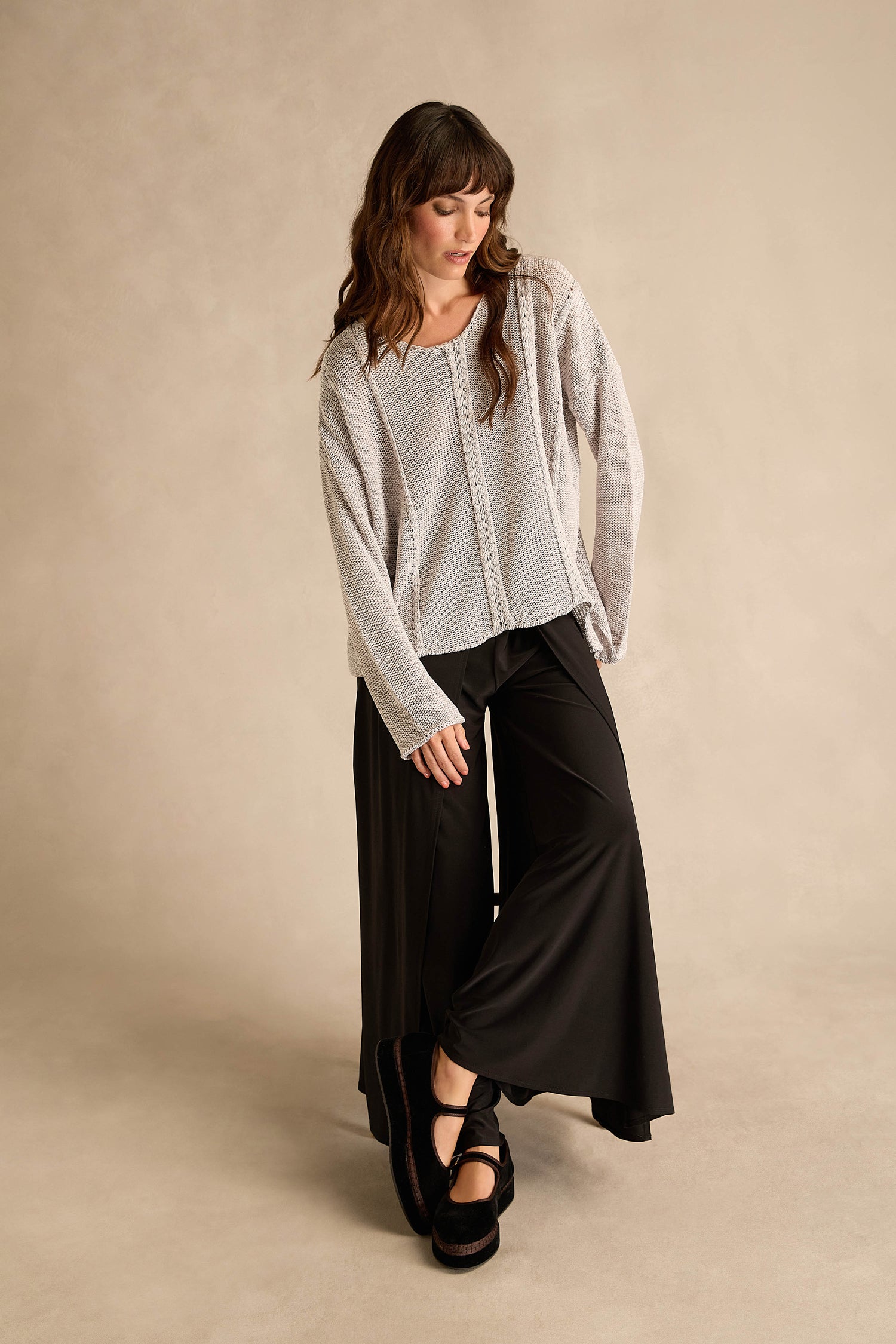 A woman stands against a beige backdrop wearing a loose gray sweater, Wide Leg Wrap Jersey Trousers in black, and black platform shoes, looking down with her arms relaxed.
