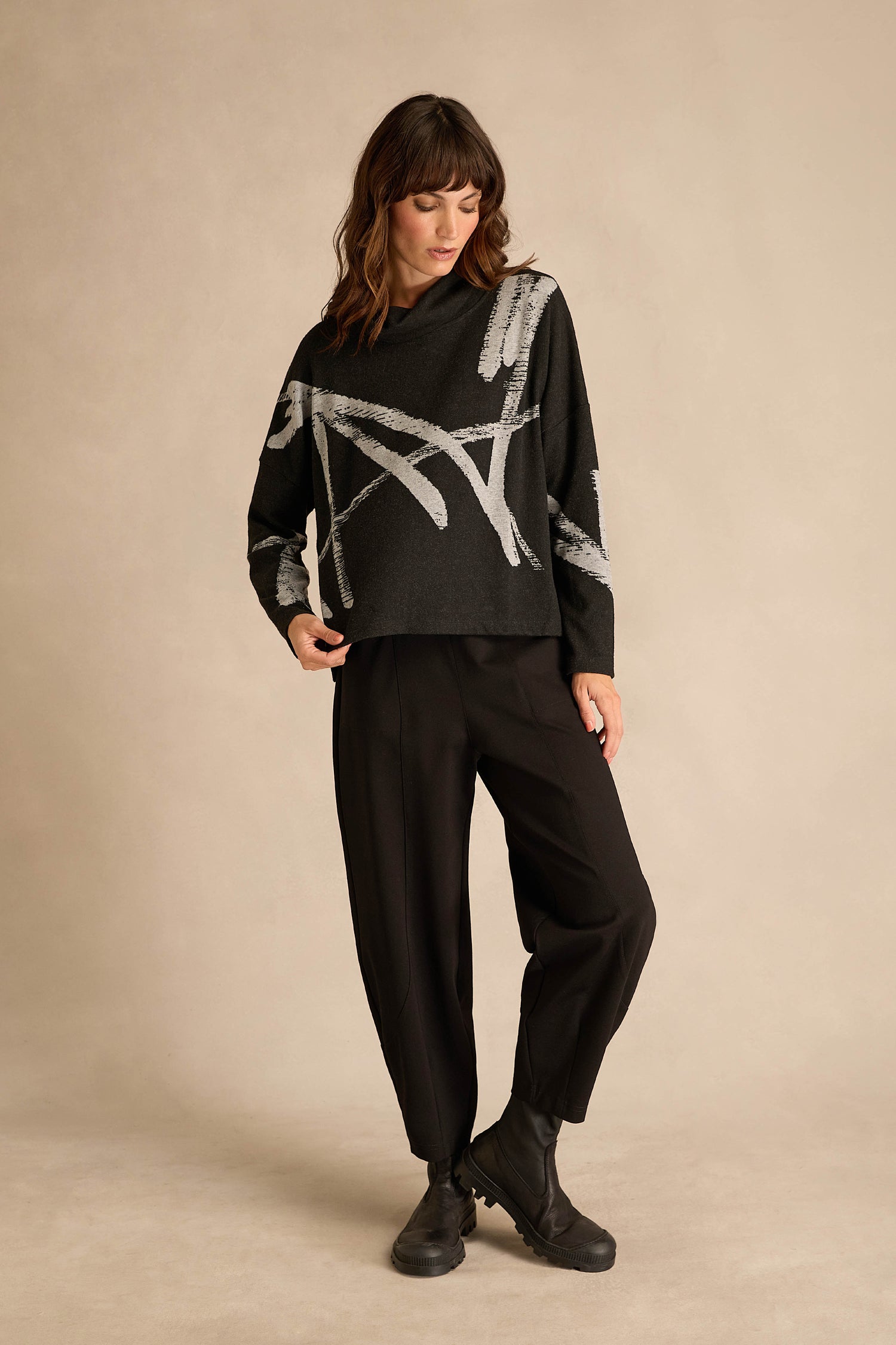 A woman stands against a beige background, wearing the Paint Stripes Long Sleeve Top— a black sweater featuring gray abstract patterns in a relaxed fit—paired with black pants and black ankle boots.