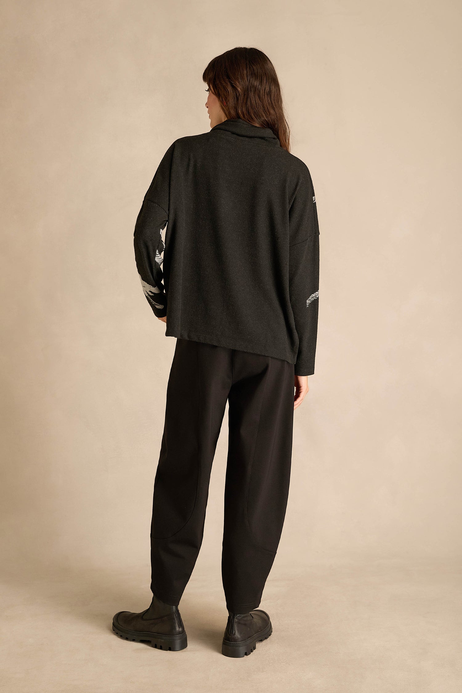 A person stands with their back to the camera, wearing the Paint Stripes Long Sleeve Top—featuring a relaxed fit and silver stripe detail—paired with loose black pants and black shoes, set against a plain beige background.