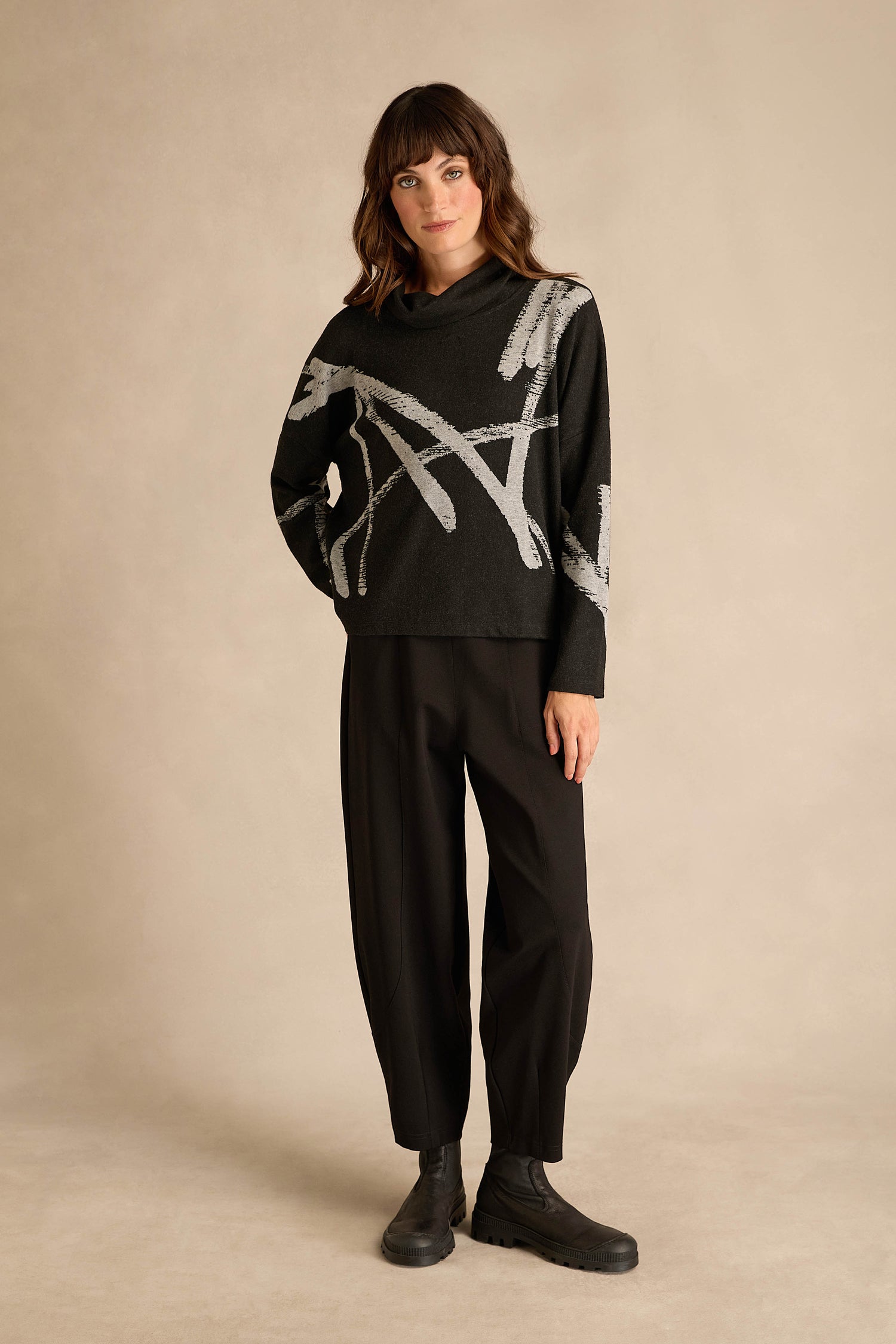 A woman stands against a beige background wearing the Paint Stripes Long Sleeve Top—a black relaxed-fit sweater with gray abstract lines—paired with black loose pants and black boots.