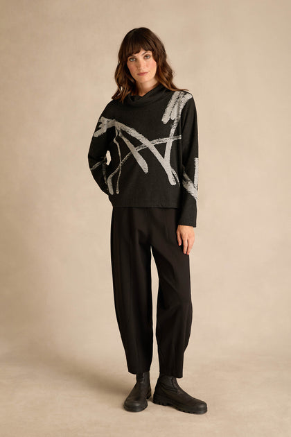 A woman stands against a beige background wearing the Paint Stripes Long Sleeve Top—a black relaxed-fit sweater with gray abstract lines—paired with black loose pants and black boots.