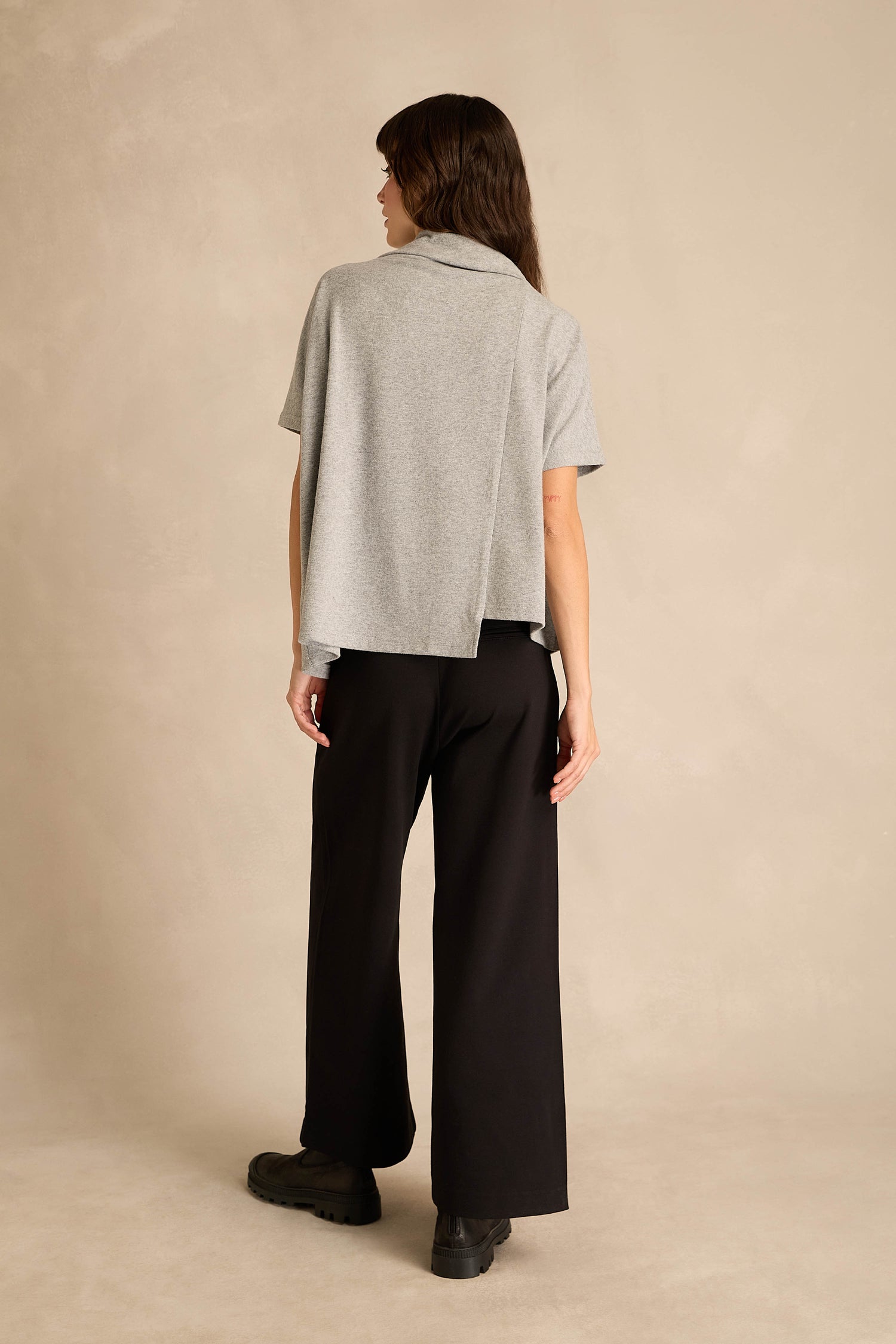 A person stands on a neutral background, facing away, wearing the Paint Stripes Draped Neck Top in gray, paired with black wide-leg pants and black shoes.