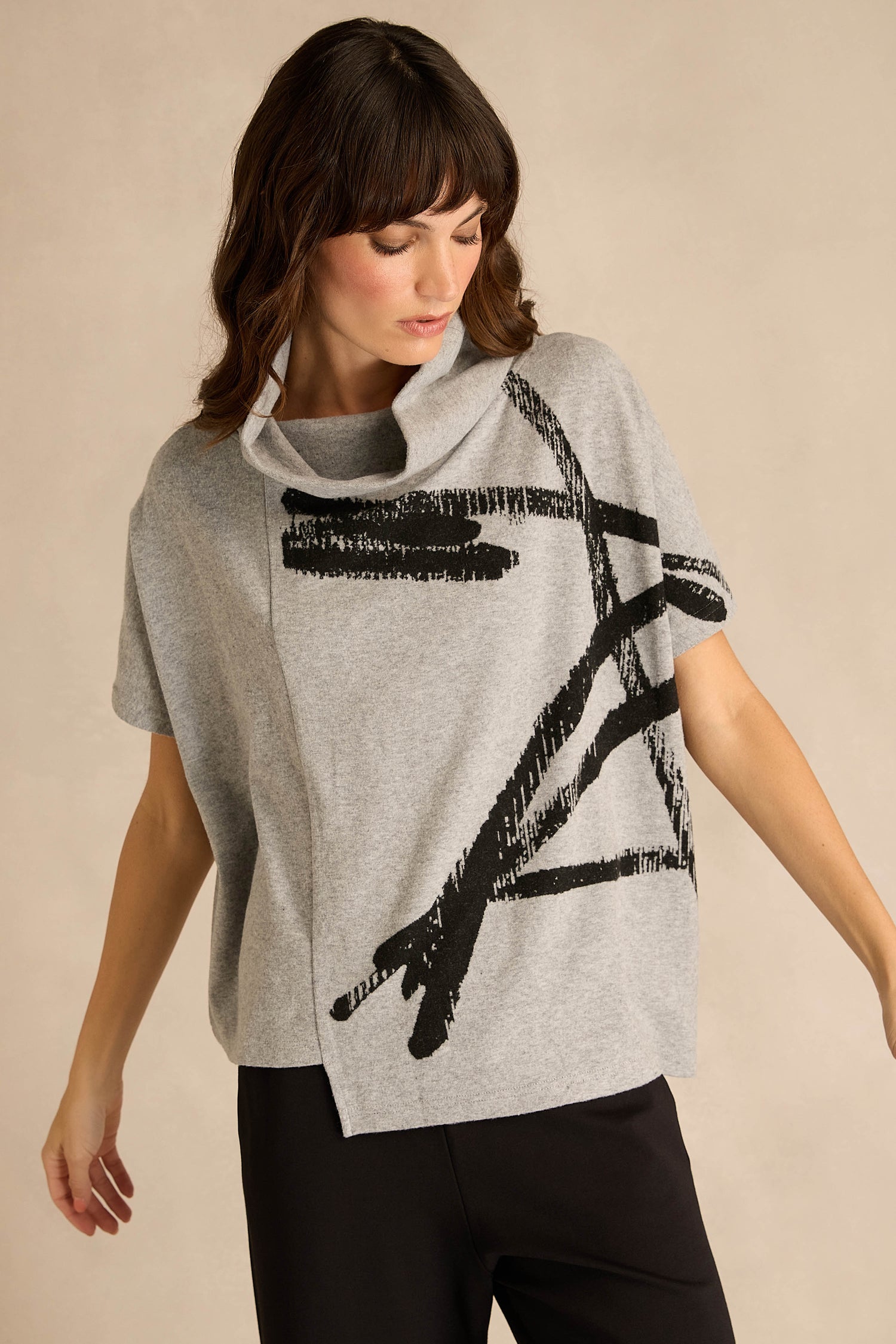 Wearing the Paint Stripes Draped Neck Top—a loose gray short-sleeved style with a black brushstroke motif—paired with black pants, a person stands against a beige background.