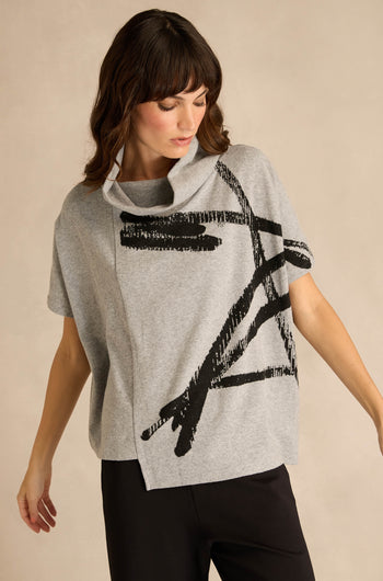 Wearing the Paint Stripes Draped Neck Top—a loose gray short-sleeved style with a black brushstroke motif—paired with black pants, a person stands against a beige background.