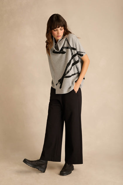 A person stands on a neutral background wearing the Paint Stripes Draped Neck Top with black abstract lines, wide black pants, and black boots.