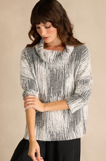 A woman wearing the Greyscale Textured Jersey Top—a loose, black-and-white jersey with a subtle textured pattern, long sleeves, and a draped neck—stands against a plain beige background, looking down and touching her arm.