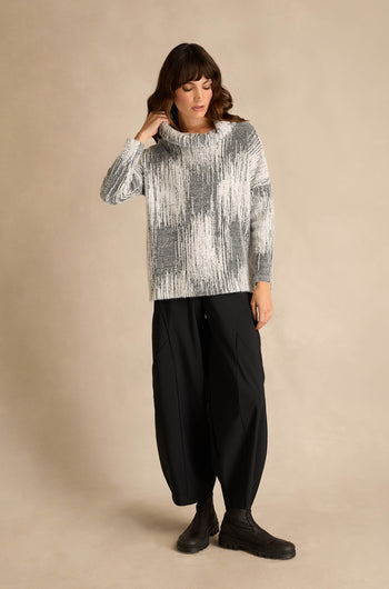 A woman wearing the Greyscale Textured Jersey Top—with its gray and white pattern and subtle textured feel—paired with black wide-leg pants and black boots, stands against a plain beige background.