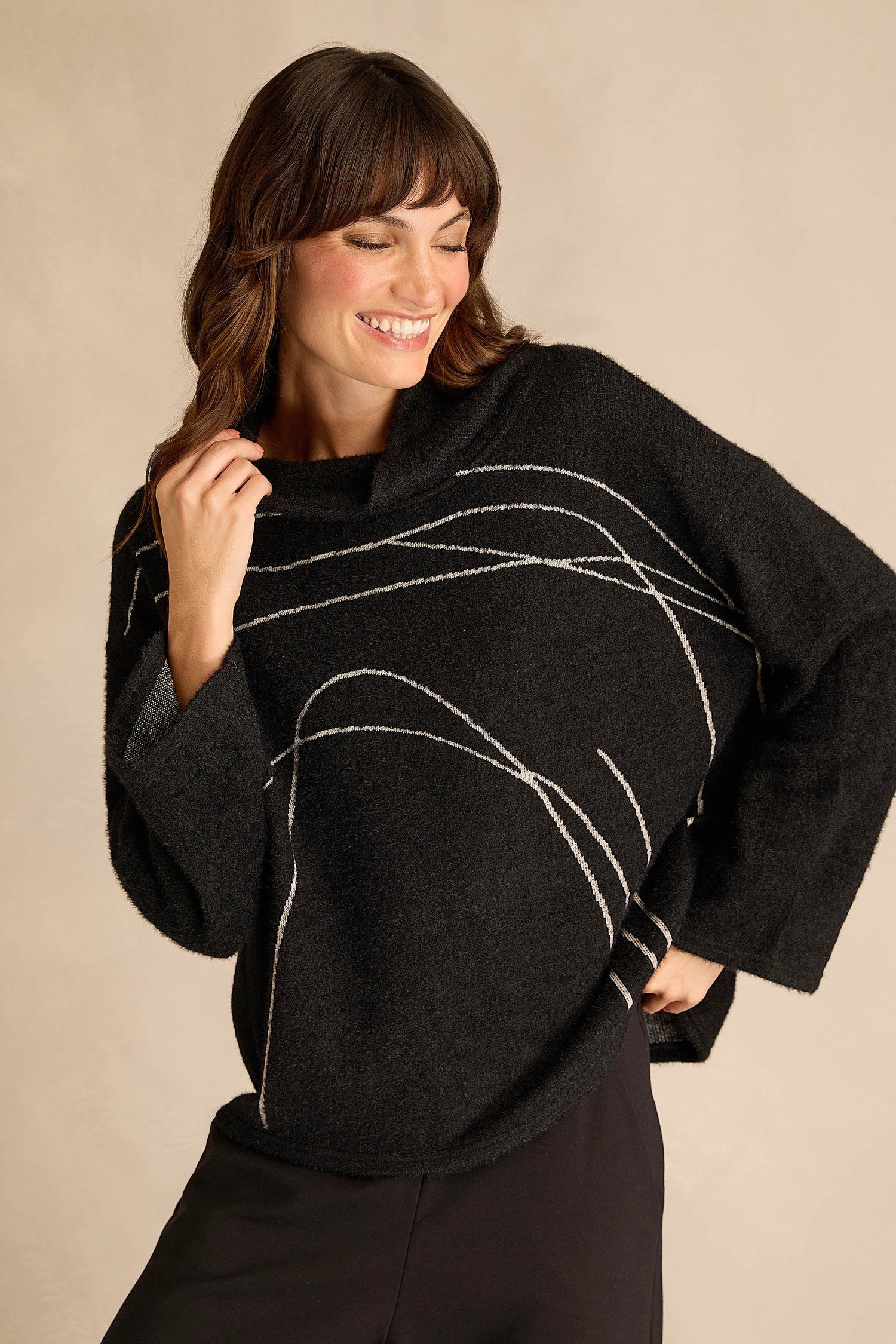 Woman smiling and posing in the Silver Lines Cowl Neck Top, a relaxed fit black sweater with white abstract lines, standing against a plain beige background.