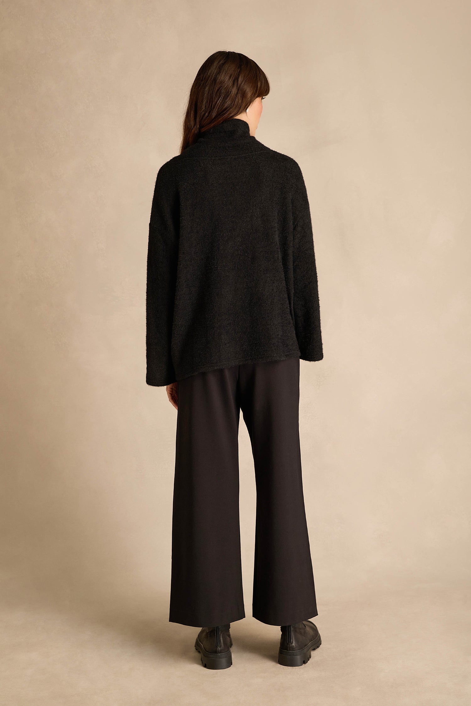 A person stands facing away against a neutral background, wearing the Silver Lines Cowl Neck Top in black, paired with wide-leg black pants and black shoes.