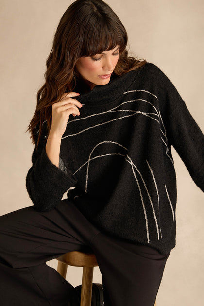 A woman with long brown hair sits on a wooden stool, looking down with one hand near her face, wearing the relaxed fit Silver Lines Cowl Neck Top—featuring white abstract lines on black—and black pants.