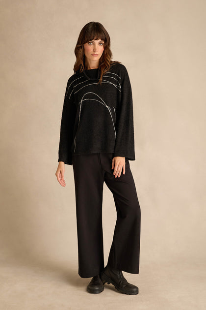 A woman stands against a beige background wearing the Silver Lines Cowl Neck Top, a relaxed fit black sweater with a silver line motif, paired with black wide-leg pants and black boots.