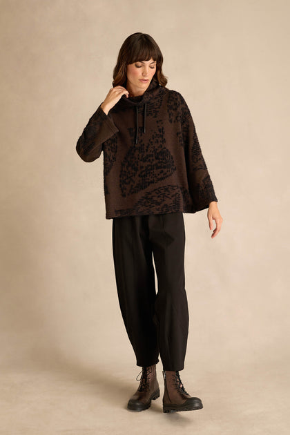 A woman stands against a beige background wearing the Cocoa Speckled Jersey Boxy Top, black loose-fitting pants, and black lace-up boots.