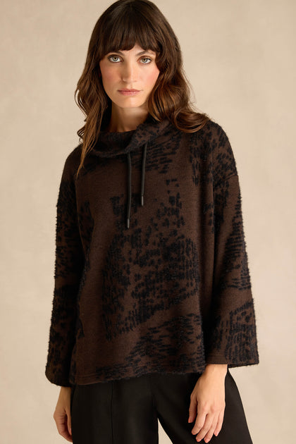A woman wearing the Cocoa Speckled Jersey Boxy Top—a loose, long-sleeve, dark brown boxy fit top with a cowl neck, black abstract pattern, and black drawstrings—paired with black pants.