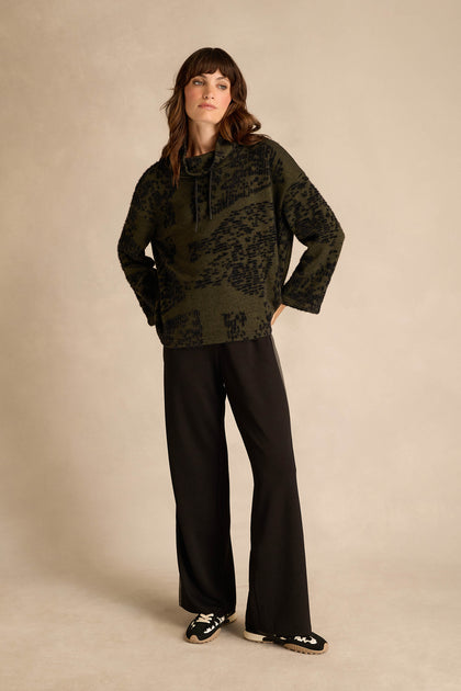 A woman stands against a neutral background, wearing the Thyme Speckled Jersey Boxy Top in green and black with a striking pattern, paired with black wide-leg pants and black slip-on shoes featuring white accents.
