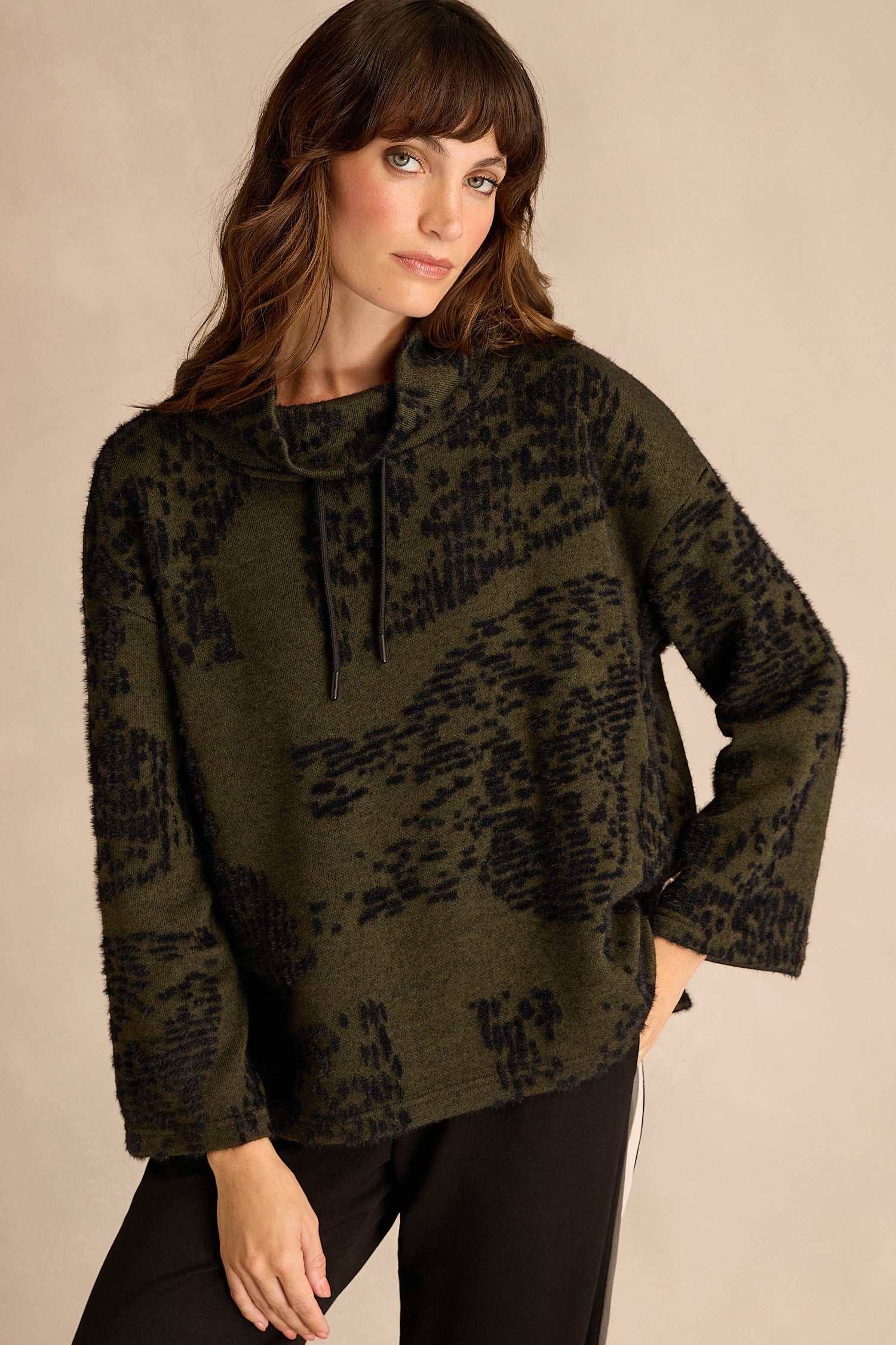 A woman stands against a neutral background, wearing the Thyme Speckled Jersey Boxy Top—an olive green and black patterned, boxy-fit long-sleeve with a cowl neck and drawstrings—paired with black pants.