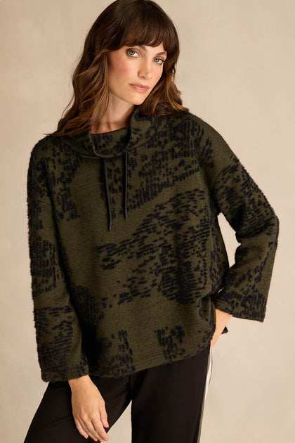 A woman stands against a neutral background, wearing the Thyme Speckled Jersey Boxy Top—an olive green and black patterned, boxy-fit long-sleeve with a cowl neck and drawstrings—paired with black pants.