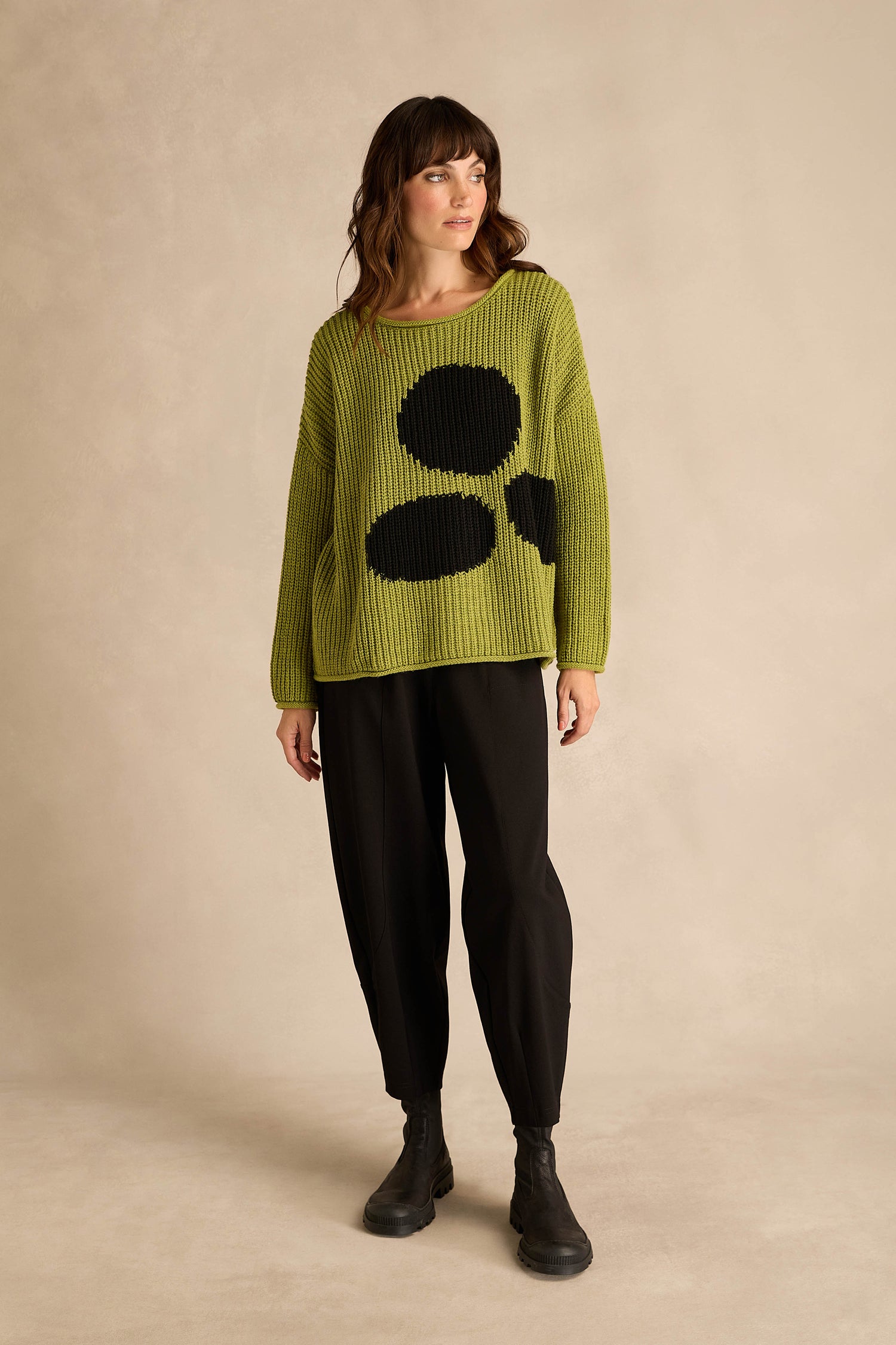 A woman stands against a neutral background, wearing the Avocado Spot Scoop Neck Knit with black pants and black boots.