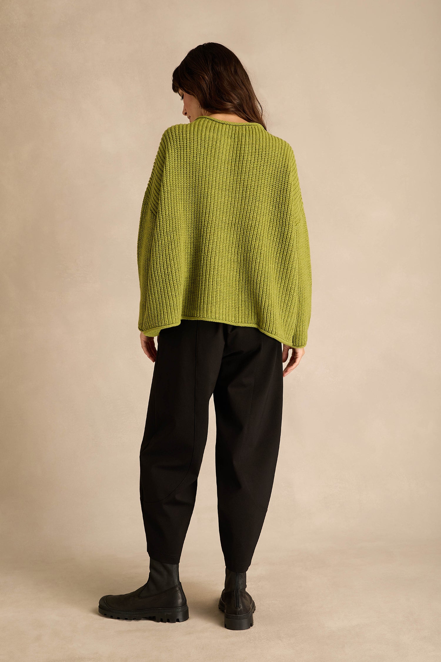 A person stands facing away, wearing the Avocado Spot Scoop Neck Knit—a loose wool alpaca sweater in green—paired with black pants and black boots, against a neutral beige background.