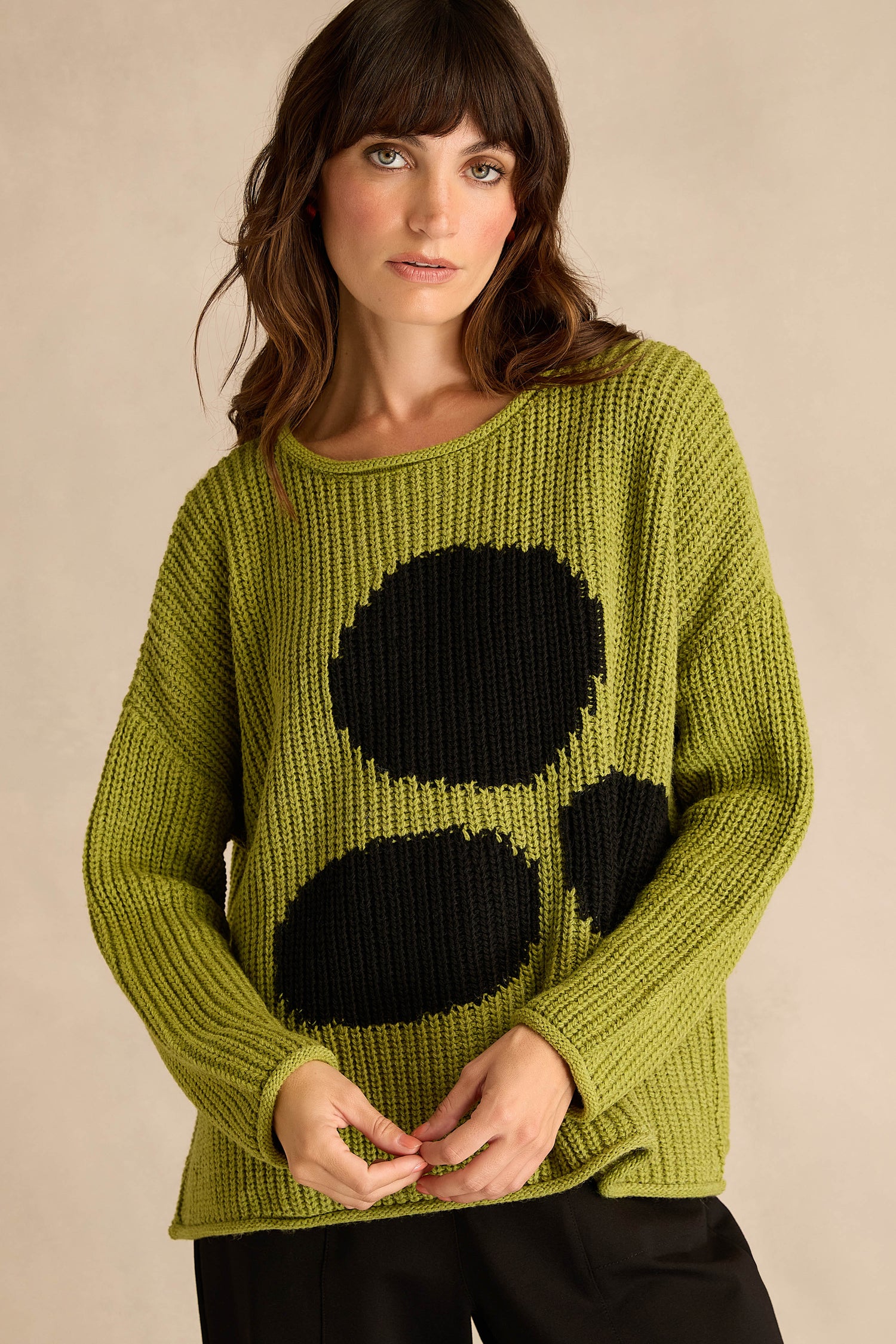 A woman wears the Avocado Spot Scoop Neck Knit—a loose-fitting green sweater featuring three large black oval spots—styled as a tunic, standing against a plain beige background.