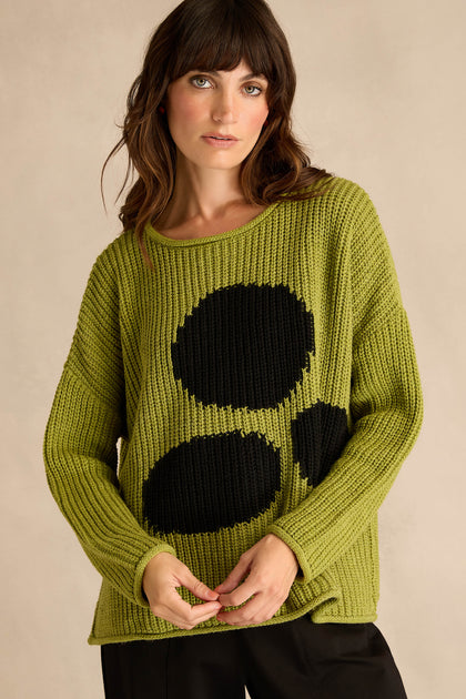 A woman wears the Avocado Spot Scoop Neck Knit—a loose-fitting green sweater featuring three large black oval spots—styled as a tunic, standing against a plain beige background.