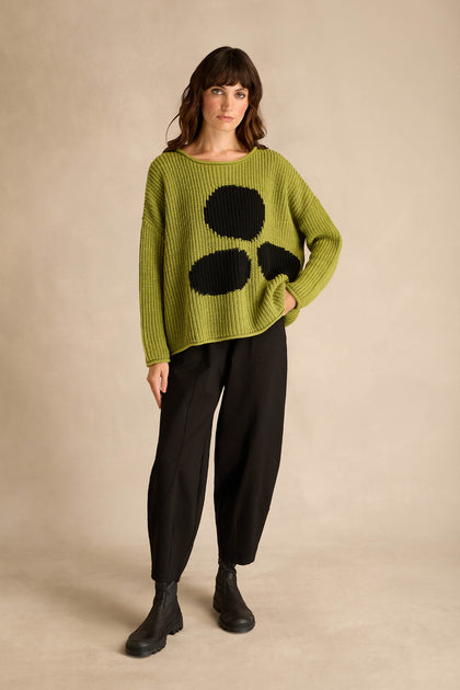 A woman stands against a plain background wearing the Avocado Spot Scoop Neck Knit, a loose green sweater with black circular designs in a cozy wool alpaca blend, paired with black pants and black boots.