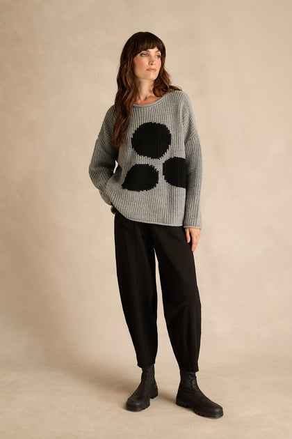 A woman stands against a neutral background, dressed in the Grey Spot Scoop Neck Knit—an alpaca and wool sweater featuring an oversized spot motif with large black circles—paired with black pants and black boots.