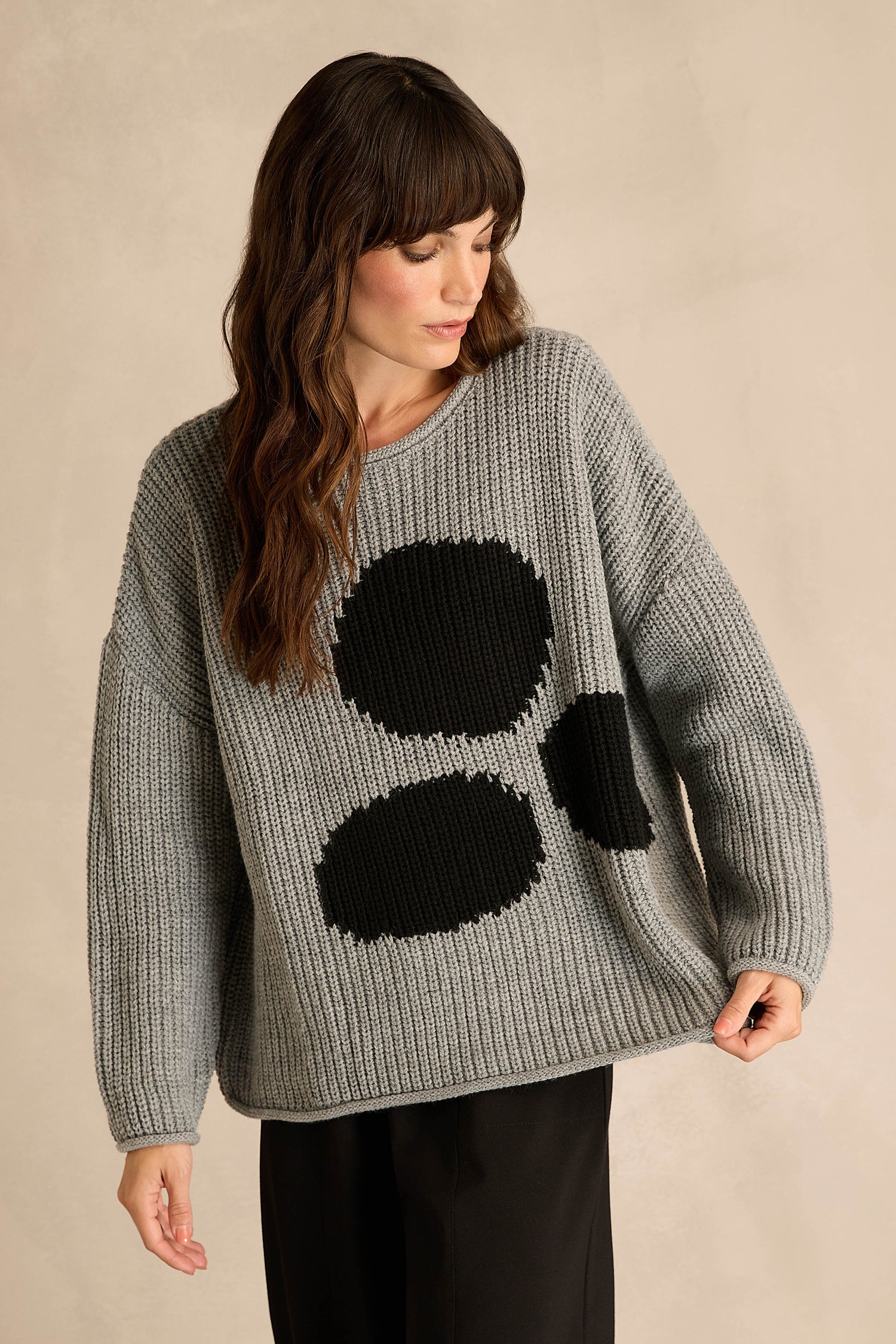 Wearing the Grey Spot Scoop Neck Knit—a loose gray sweater with an oversized spot motif featuring three large black dots on the front, crafted from a soft wool and alpaca blend—styled with black pants against a plain beige background.