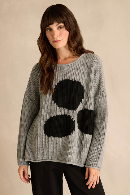 A woman wears the Grey Spot Scoop Neck Knit—a gray oversized sweater featuring a spot motif, crafted from a cozy wool and alpaca blend with a relaxed scoop neckline—paired with black pants and set against a beige background.