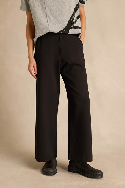 A person stands against a neutral background wearing a grey short-sleeve shirt, the Wide Leg Ponte Trousers in black, and black shoes.