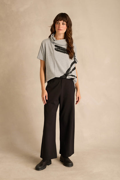 A person stands against a beige background wearing a gray short-sleeve top with black abstract lines, the Wide Leg Ponte Trousers in black crafted from stretch ponte jersey, and black shoes.