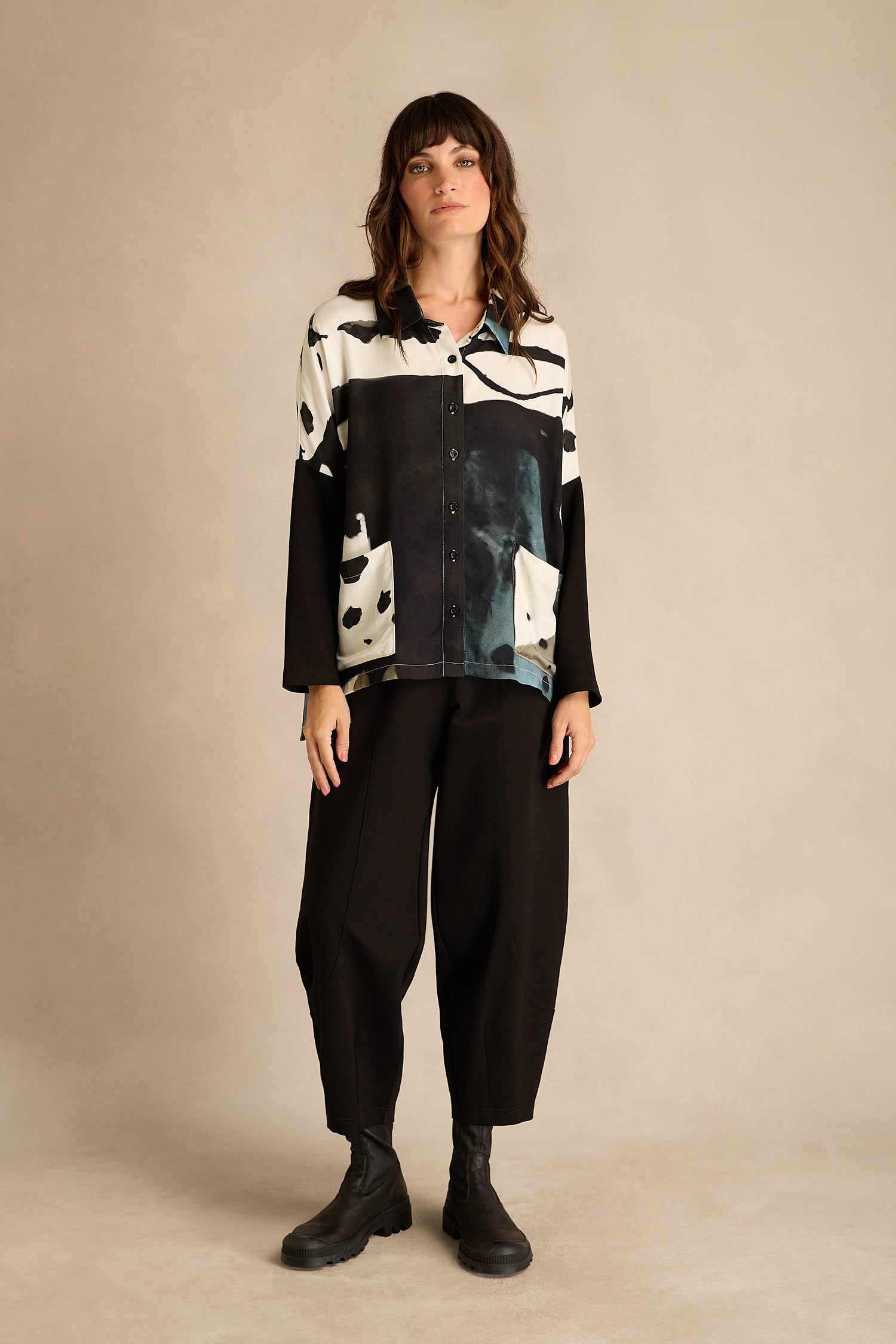 A woman stands against a neutral background wearing the Abstract Watercolour Shirt, paired with loose black pants and black boots.