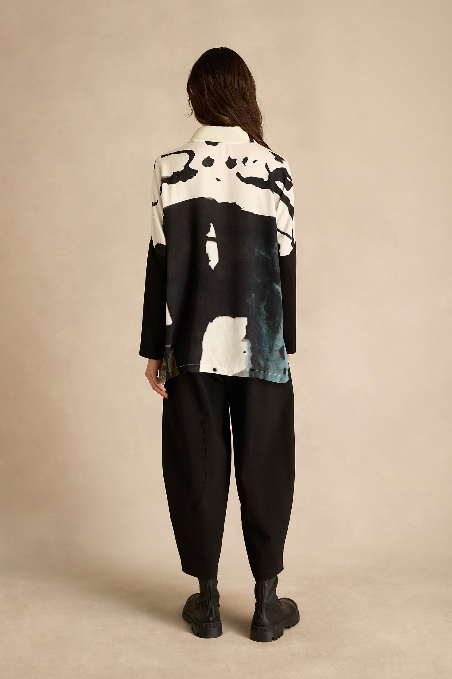 Person standing with back to camera, wearing the Abstract Watercolour Shirt, loose black pants, and black boots against a plain beige background.