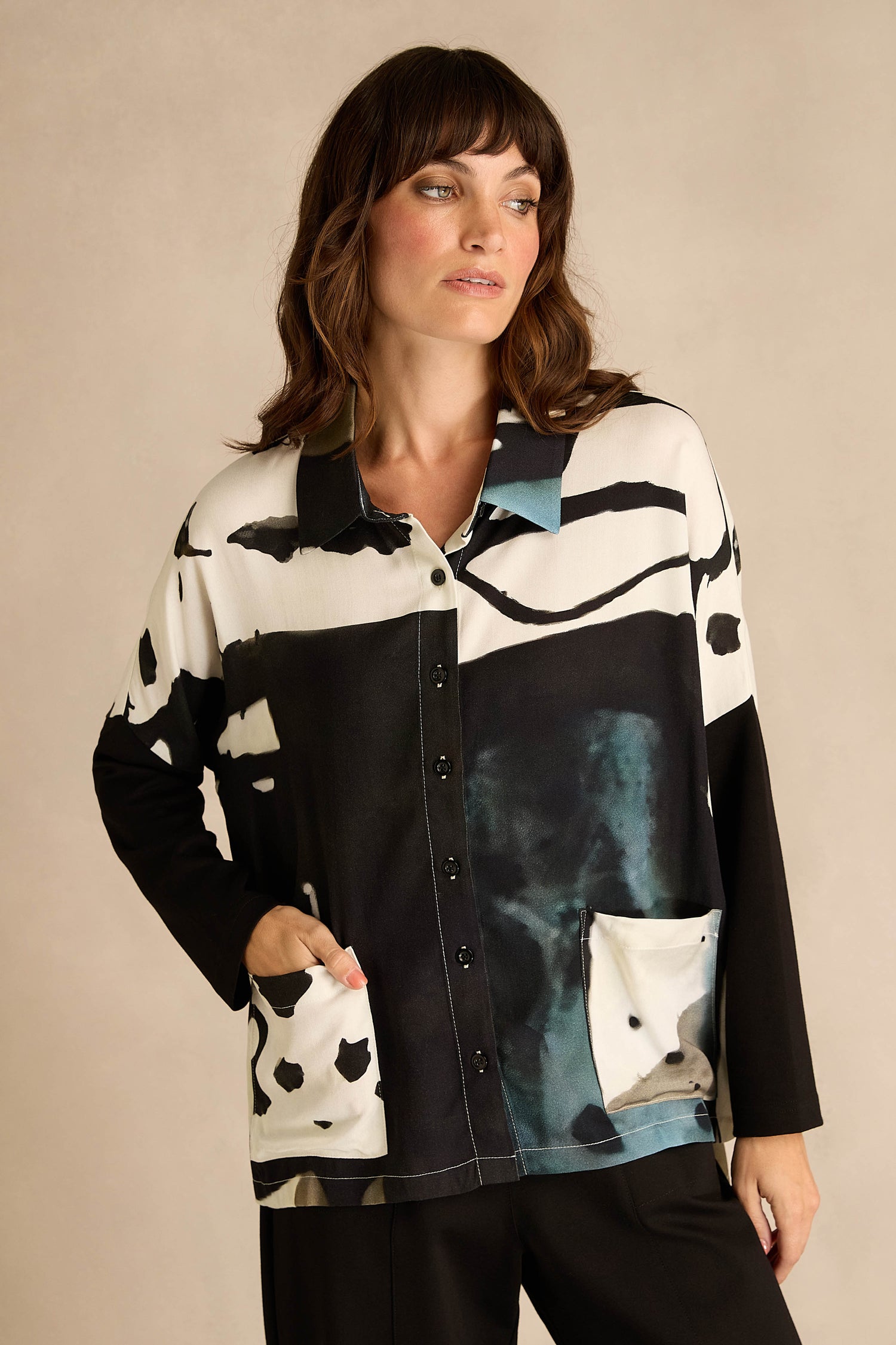 A woman wears the Abstract Watercolour Shirt, a long-sleeve, button-up top featuring black, white, and teal watercolour patterns, as she stands with one hand in her pocket against a neutral background.