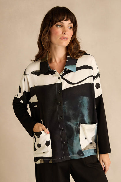 A woman wears the Abstract Watercolour Shirt, a long-sleeve, button-up top featuring black, white, and teal watercolour patterns, as she stands with one hand in her pocket against a neutral background.