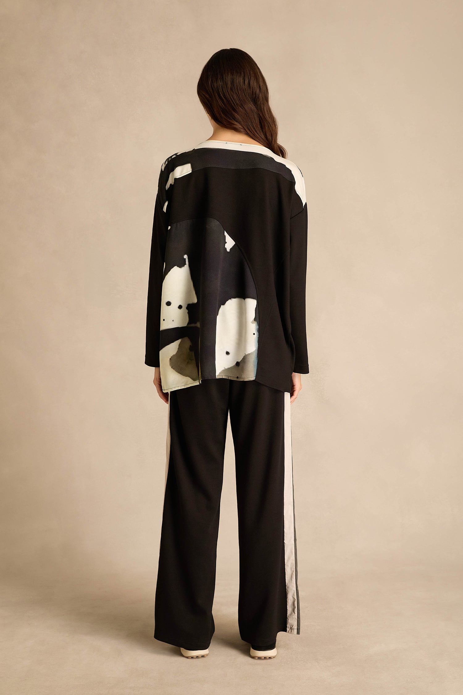 A person stands with their back to the camera, wearing loose black pants and the Abstract Watercolour Scoop Neck Top, which features a striking white abstract pattern on the back, set against a neutral background.