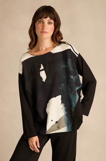 A woman stands against a neutral background, wearing the Abstract Watercolour Scoop Neck Top—a long sleeve, relaxed fit black top featuring an abstract black, white, and gray watercolor pattern—paired with coordinating black pants.
